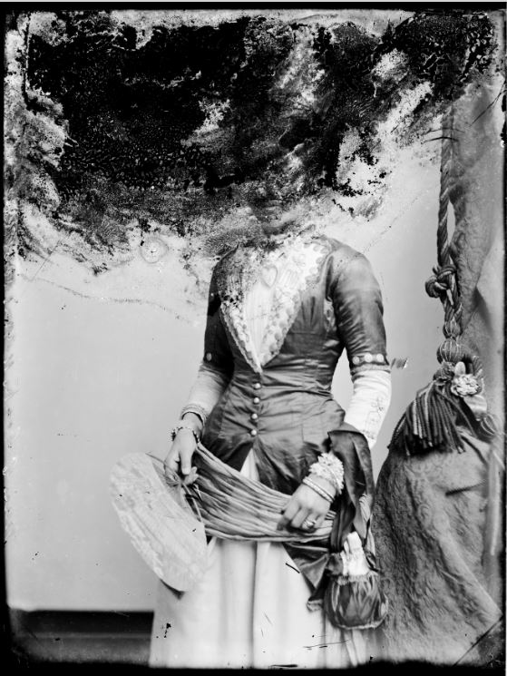 Black and white photograph of a woman in long dress, holding a fan. The image has been badly damaged at the top, almost completely obstructing her head.