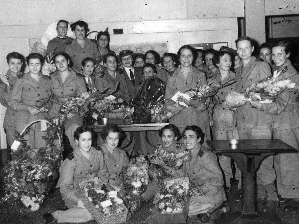 Shows around 2 women in a room. Many of them are smiling and holding bunches of flowers.