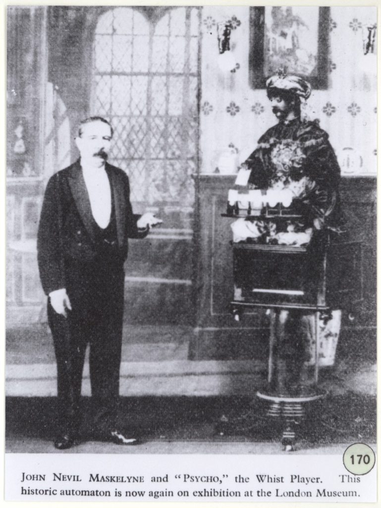 Black and white photograph of a man in a suit shown whole-length, standing next to a figure of man in eastern dress and turban, seated on a box - apparently an automaton.