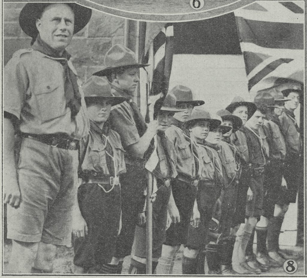 Black and white newsprint image of a man dressed in the Scouts uniform, next to twelve young boys also in Scouts uniform, standing in a line. The second boy is holding what appears to be the Australian flag.