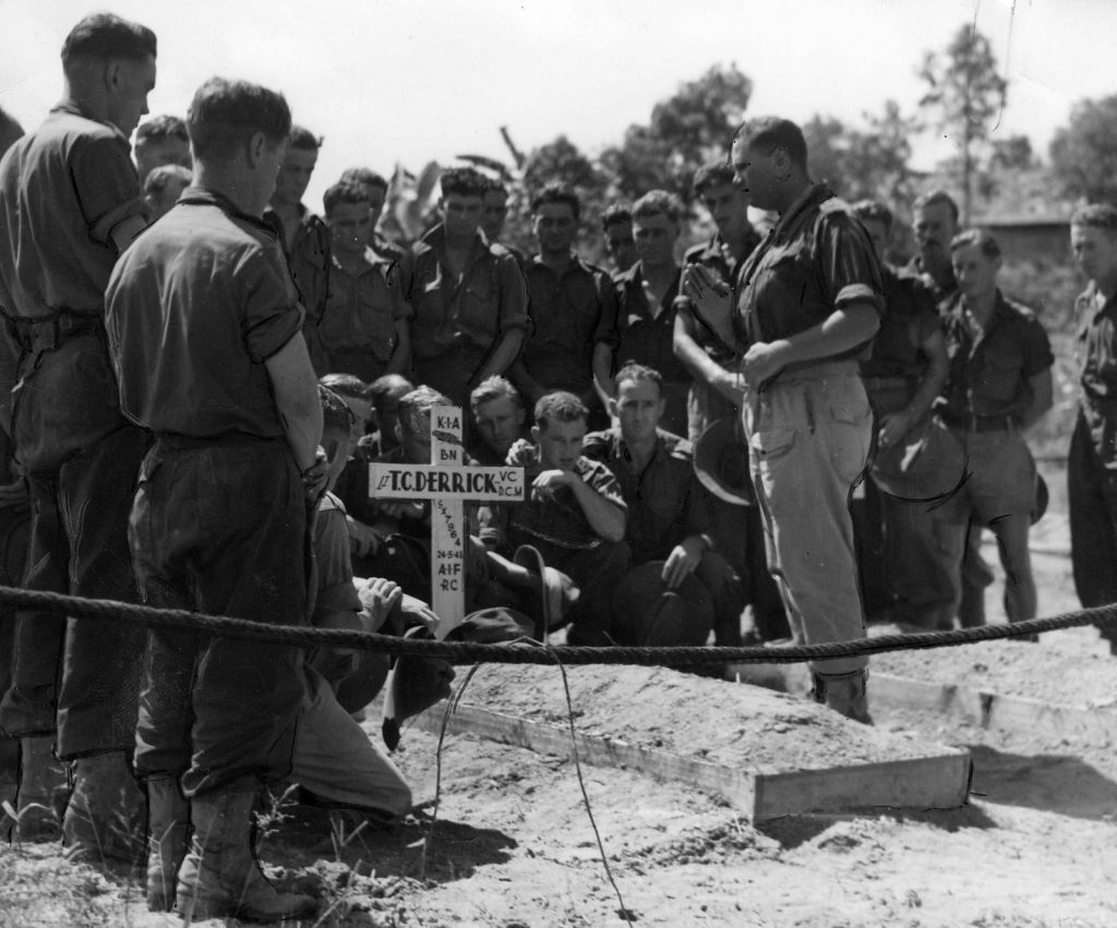 Shows the Army Padre standing over the closed grave of Lieutenant Derrick  A group of standing and squatting soldiers surround the grave.