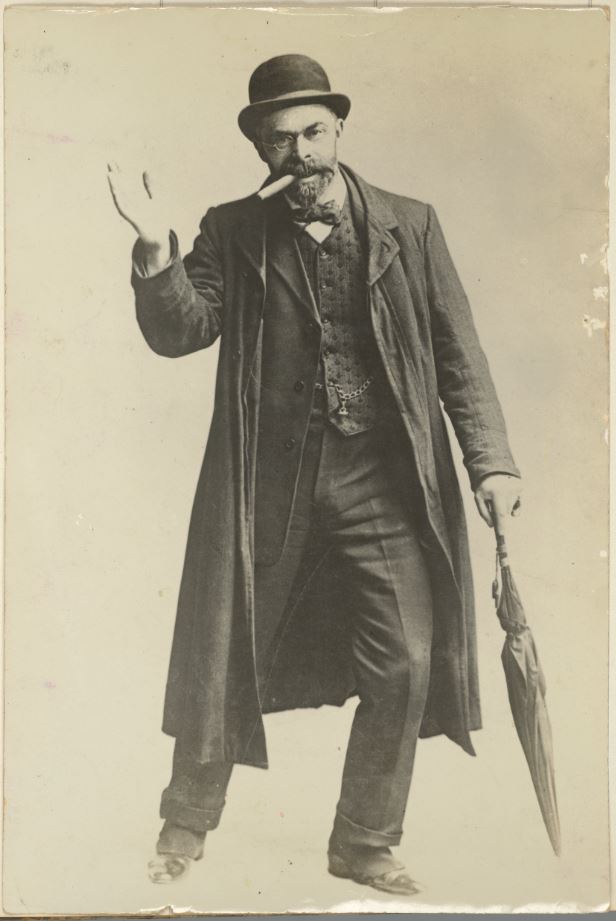 Piggott dressed up in a disguise of a London gentleman wearing a bowler hat, trench coat, waistcoat, jacket and trousers. He is carrying an umbrella and has a cigar in his mouth.