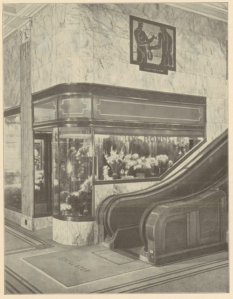 Black and white photograph on beige background showing corner of arcade with florist shop in background and the bottom of an escalator in front of it. There is an image high on the wall on the left of two dark figures. 