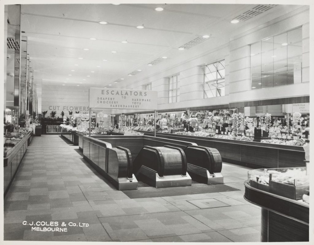 Black and white photograph showing open area with high windows on left-hand side. Around the walls are shop counters with objects on them. In the middle is the top of two escalators. A sign above the escalators reads 'Escalators to drapery crockery hardware toys haberdashery'. On the back wall a sign reads 'Cut flowers'. 