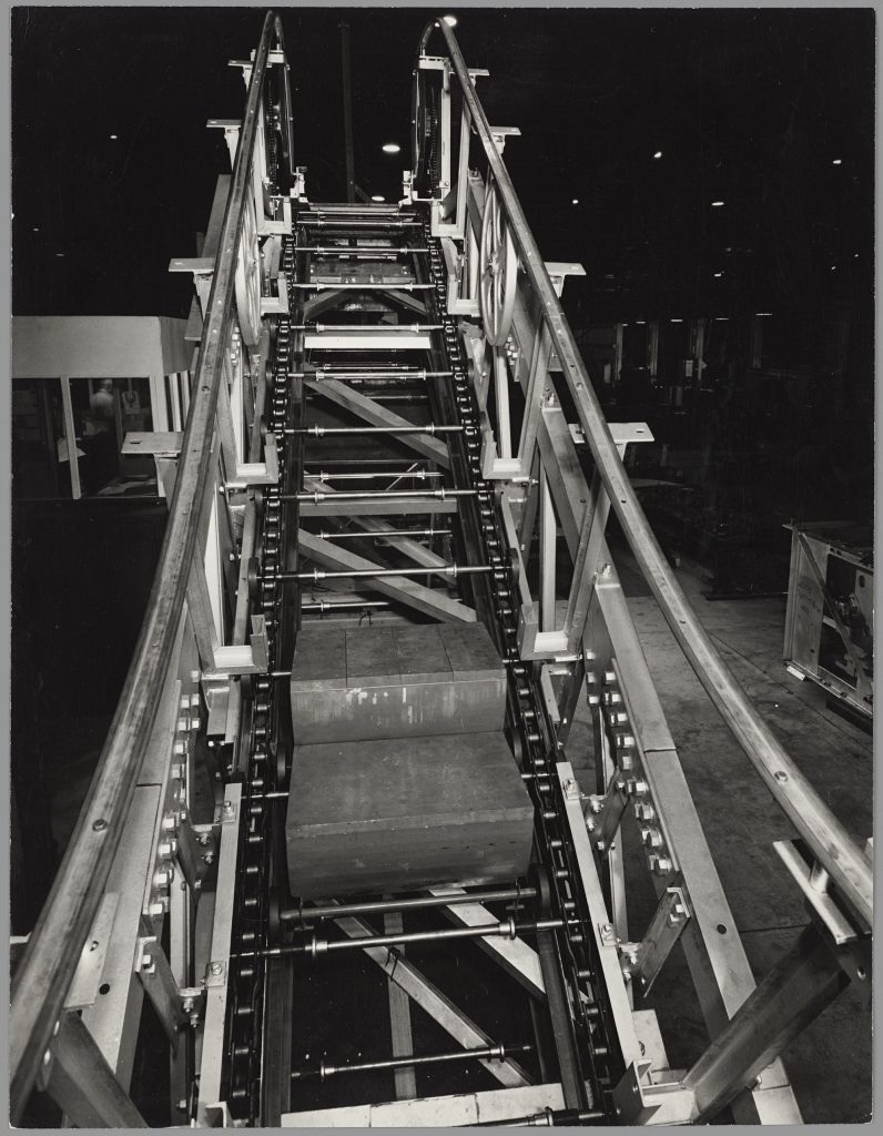 Black and white photograph showing front-on view of an escalator under construction showing metal railings either side and two rows of chains joined by bars at regular intervals. Two wooden steps are between the chains towards the bottom of the image. 