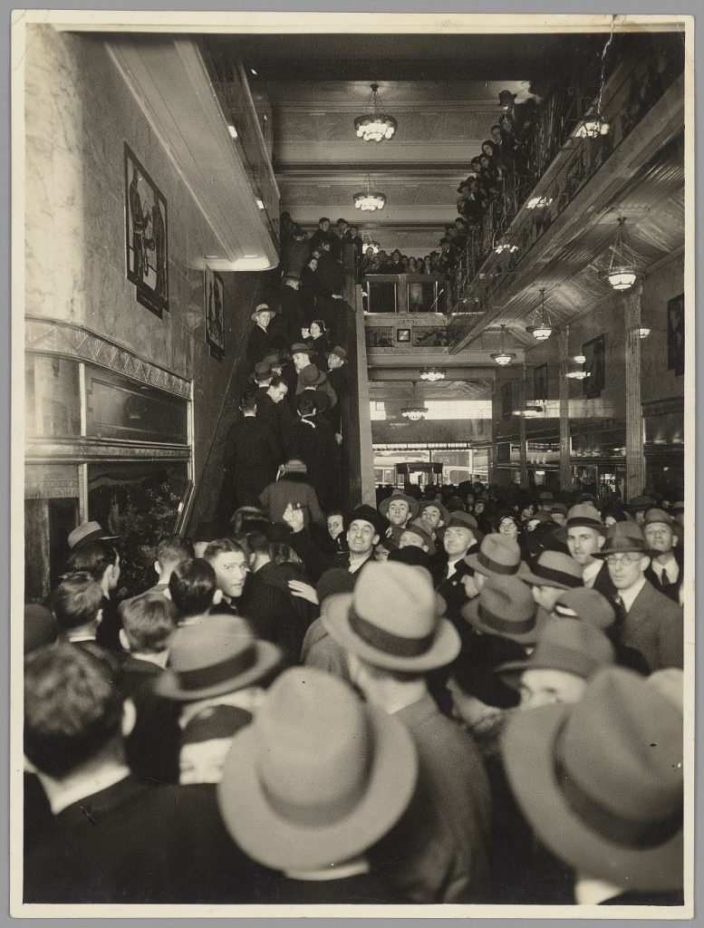 Sepia-tone photograph showing large crowd with many men in hats in an arcade, at the bottom of an escalator to the left-hand side of the arcade and some people going up the escalator and other people looking down from a balcony on the first floor that is on the right-hand side of the image. 