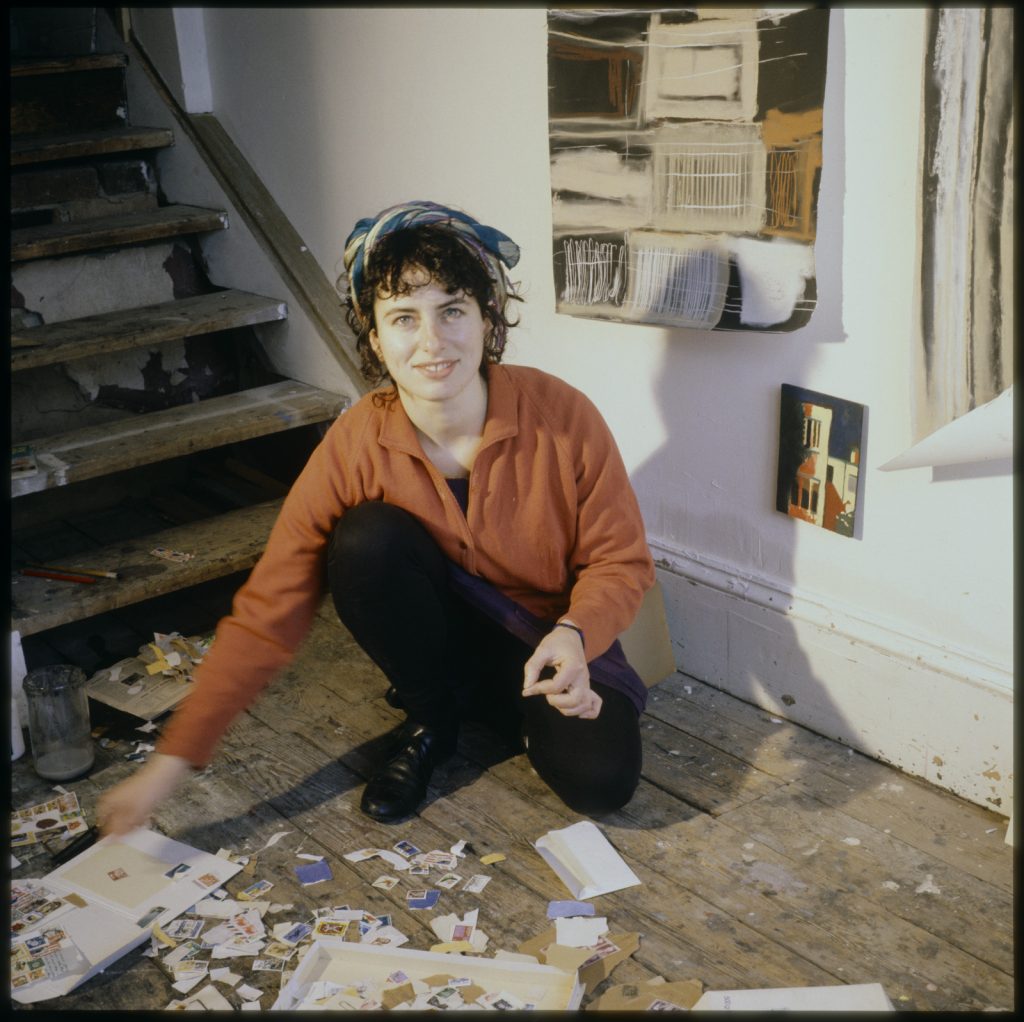 Photo of Brigid Courtney crouched down on the floor, looking at the camera and reaching out to her material in front of her.