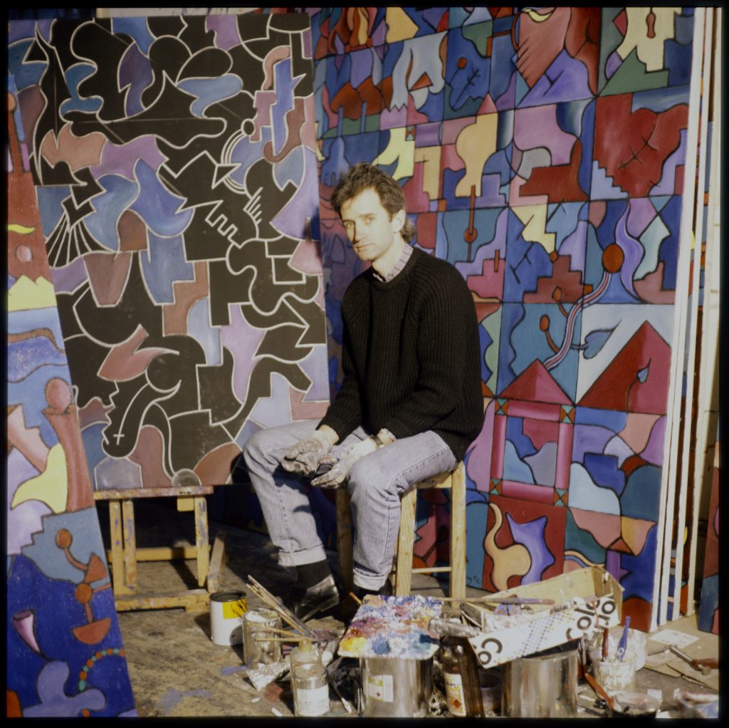 Photo of David Slattery sitting in front of his large, colourful art pieces.
