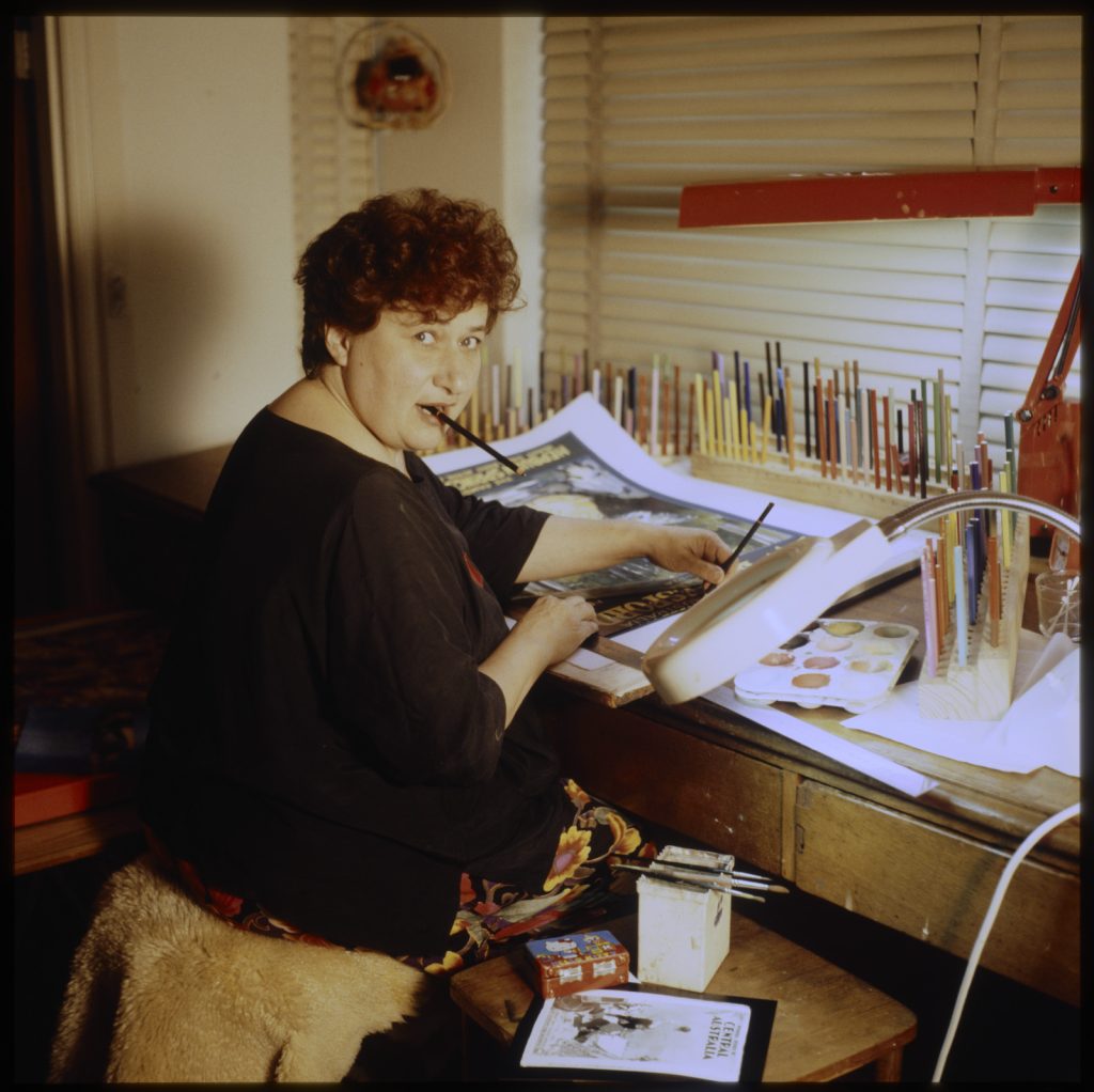Photo of Fay Plamka sitting at her desk, turned to the camera with a pencil in her mouth and another in her hand over her work.