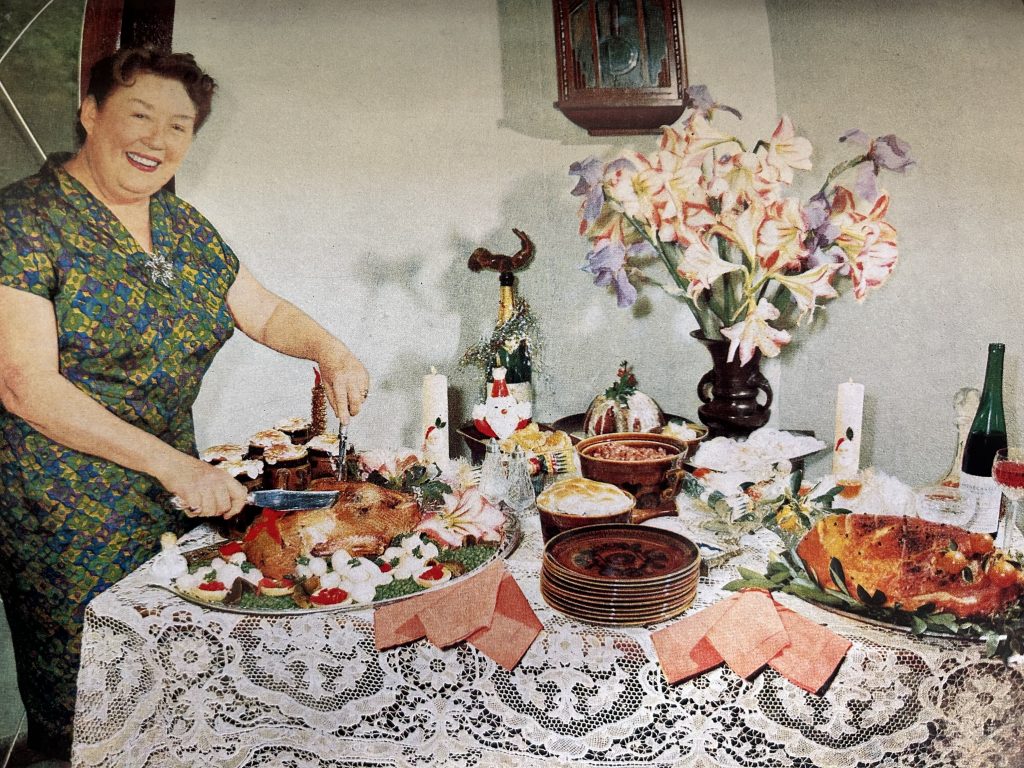 Colour photo depicting a woman carving a roast duck, with other food on decorated table