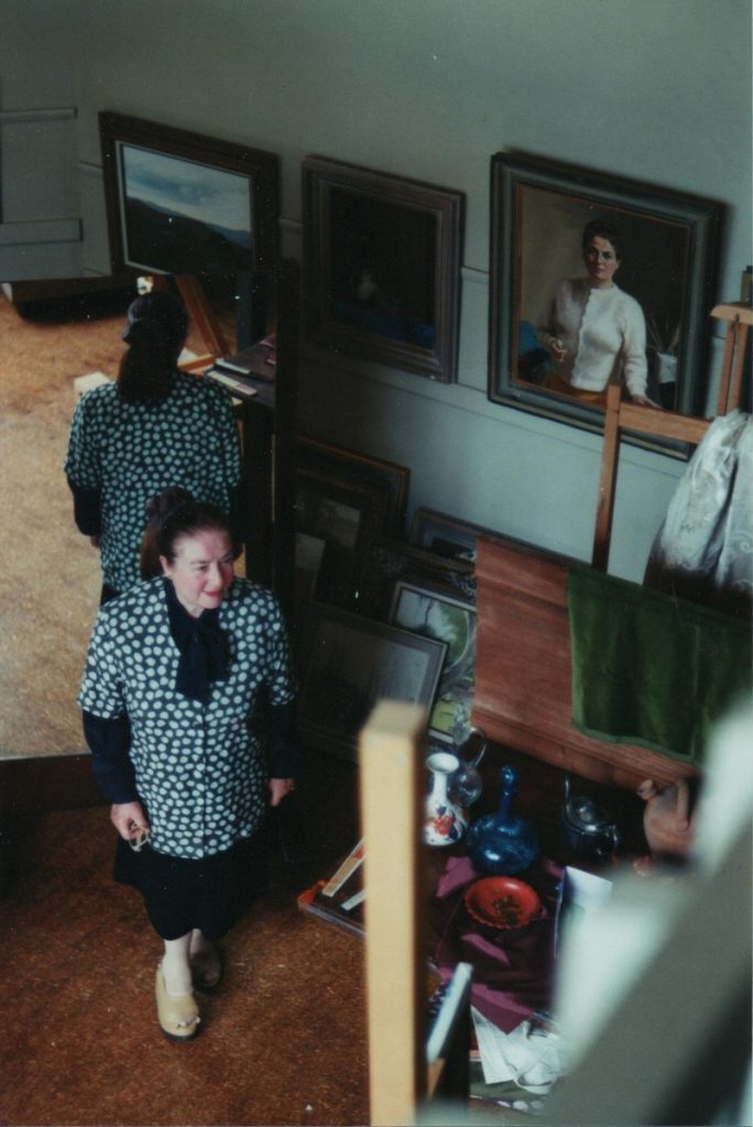 Photo from above shows JoyceMcGrath standing in her studio with her back to a mirror. Her self portrait is on the wall in the background.