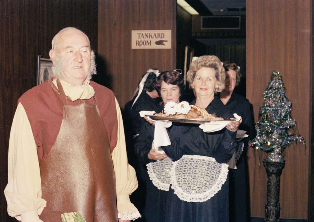 Colour photo depicting a man in an apron and women carrying platters of food.