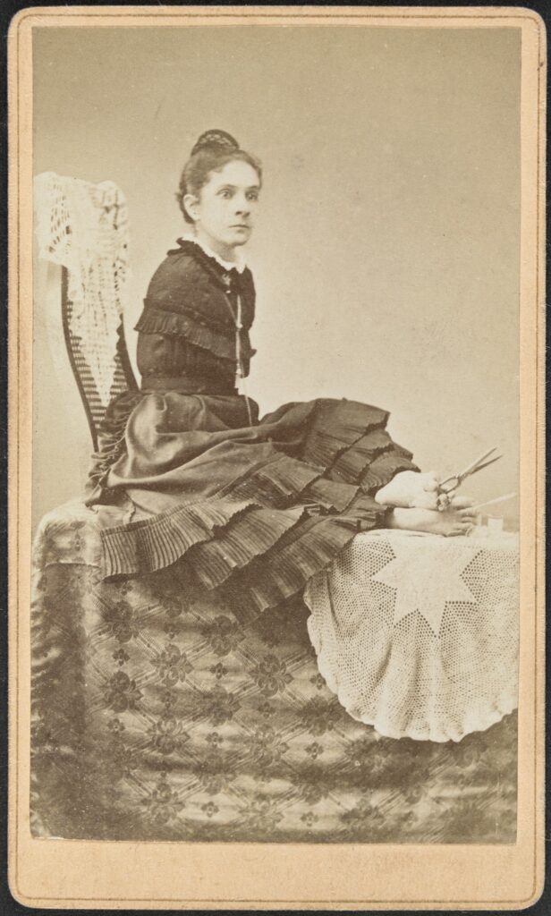Studio portrait of a woman, full length, seated, facing to her left, holding scissors and thread with her feet. It appears that she has been working on the white cloth underneath her feet.