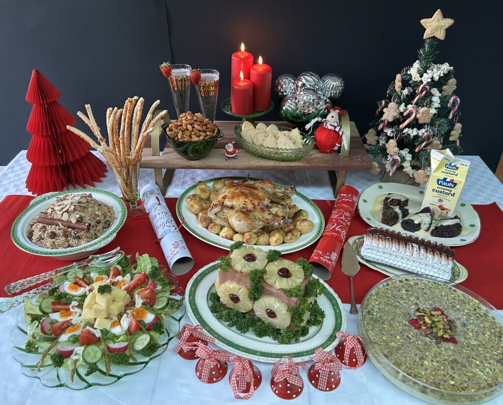 Colour photo of a decorated table holding various Christmas foods, including roast chicken, potatoes, rice, salad, pudding, glazed ham and ice cream desserts