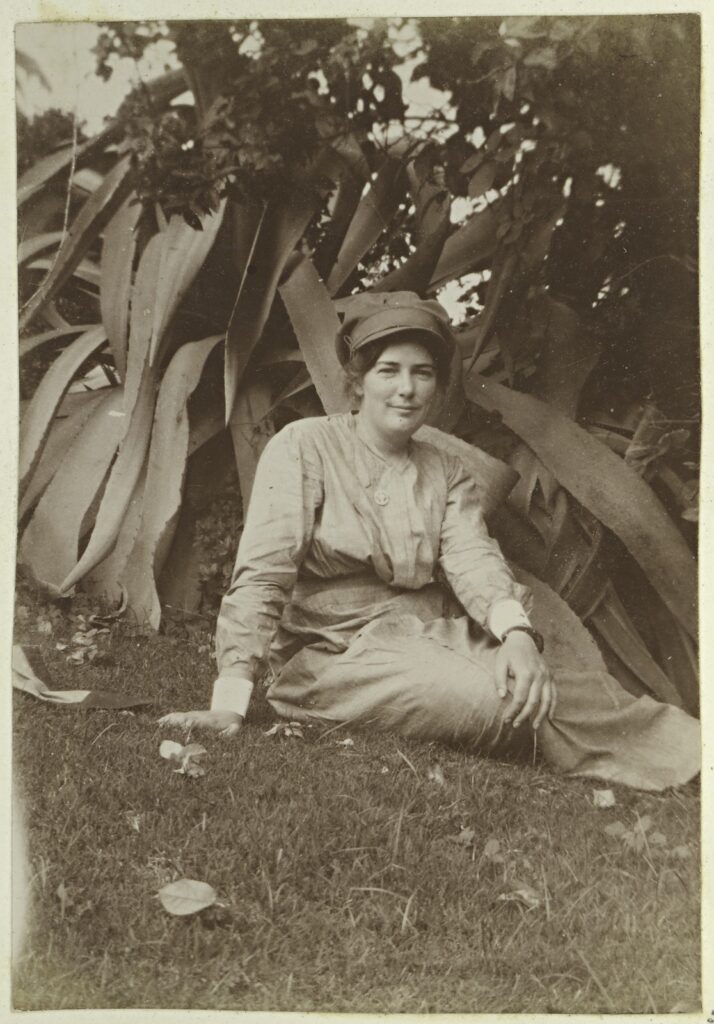 Whole-length portrait black-and-white photograph of a woman, wearing a walking out dress and military cap, sitting on a lawn.