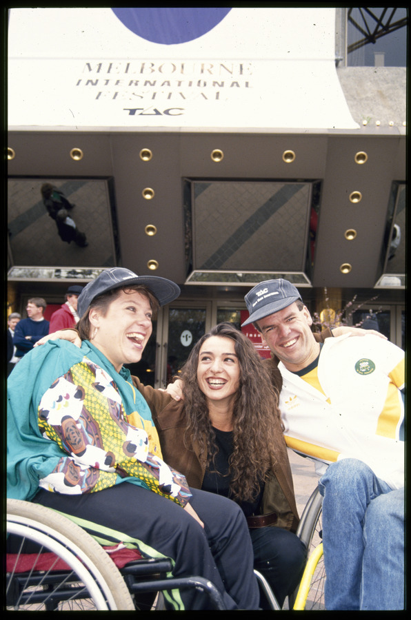 Woman and man in wheelchairs with a woman kneeling between them, outside the Arts Centre Melbourne, above signage promoting Melbourne International Festival sponsored by TAC. The man and woman in wheelchairs are wearing TAC caps. All three people have wide smiles with arms around each other. The woman in the centre is looking up at woman in the wheelchair.