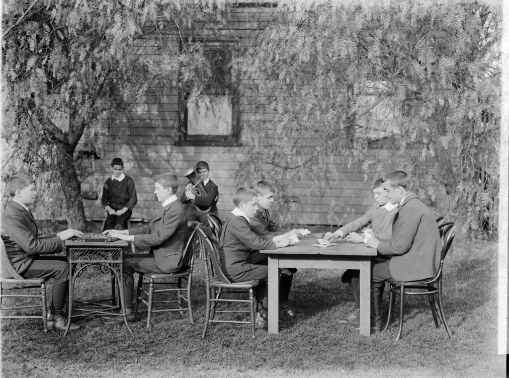 Vision-impaired boys outdoors for recreation. At left, two boys are seated across from one another playing a board game. At right, four boys are seated at a table, two on each side of the table, playing cards. A boy sits at left under a tree watching on, while next to him, a boy is playing the tuba.