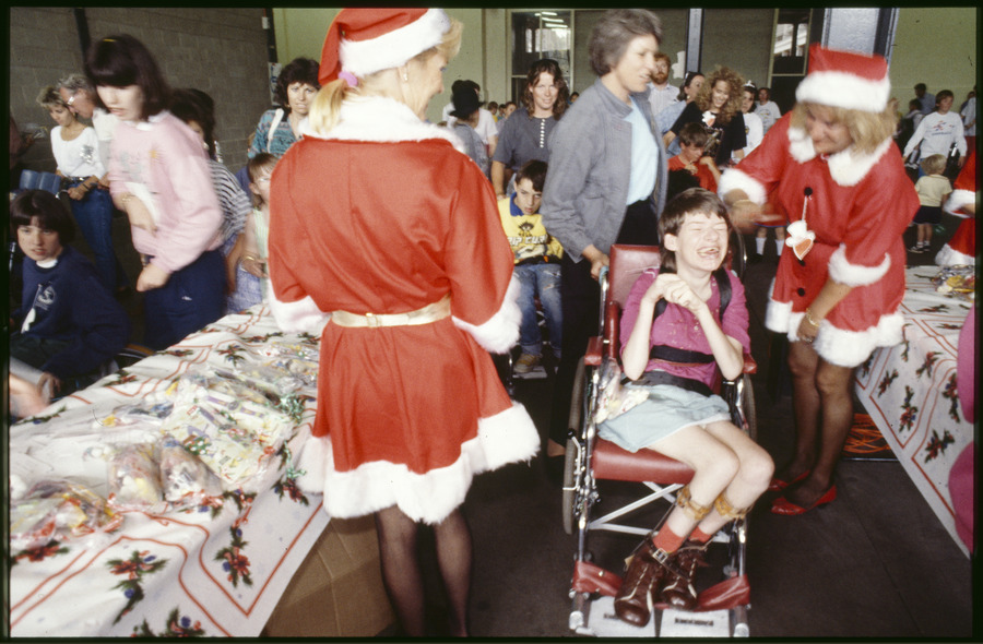 Two ladies dressed in Santa suit dresses and hats happy engaging on either side of a smiling young girl in a manual red wheelchair wearing calipers. A woman is holding onto the left handlebar of the wheelchair in side profile. The style of dress suggests the time period is the 1980s.
