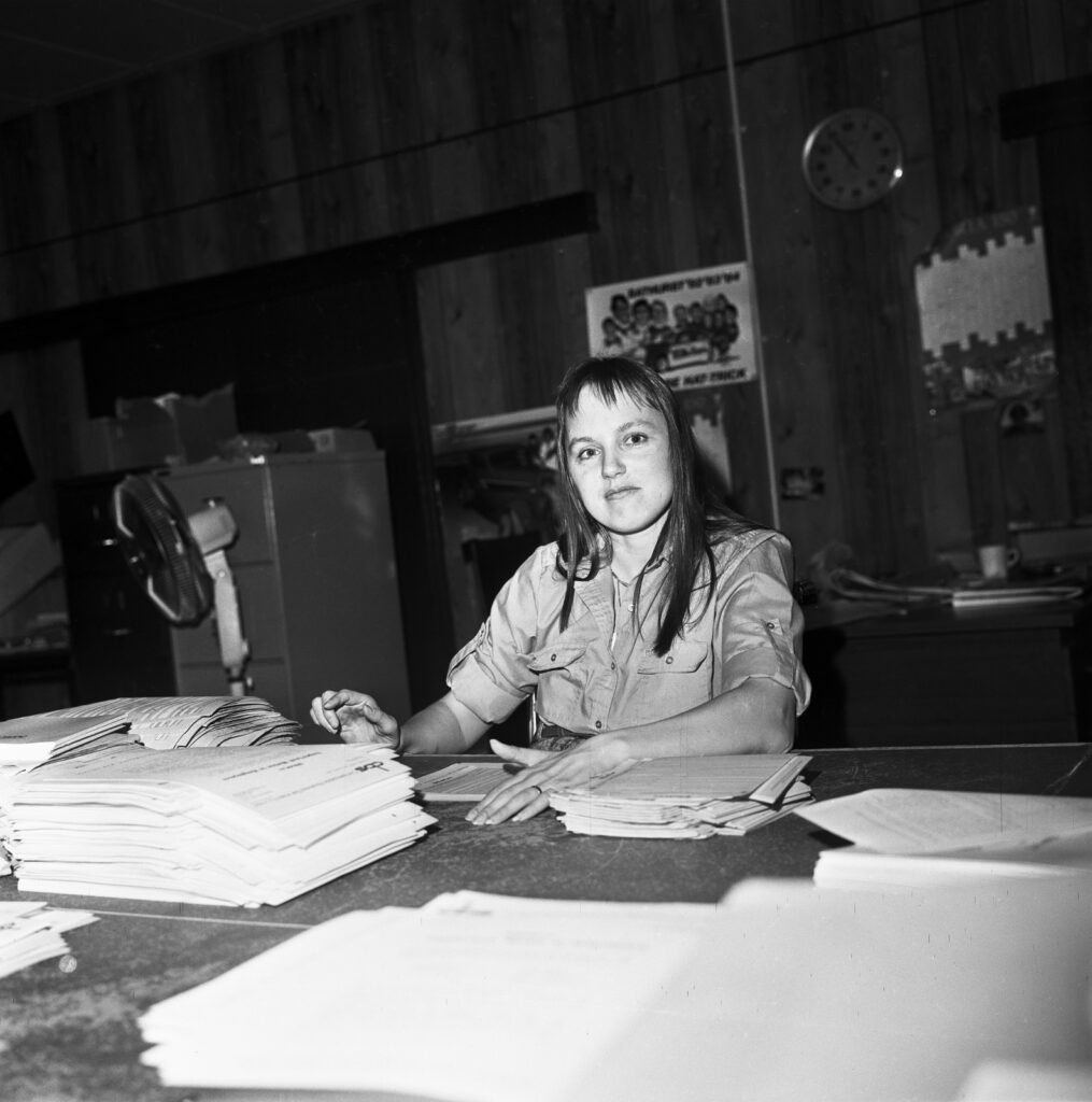 Black and white photograph of a woman sitting at a workdesk stacked neatly with papers. She is facing the camera.