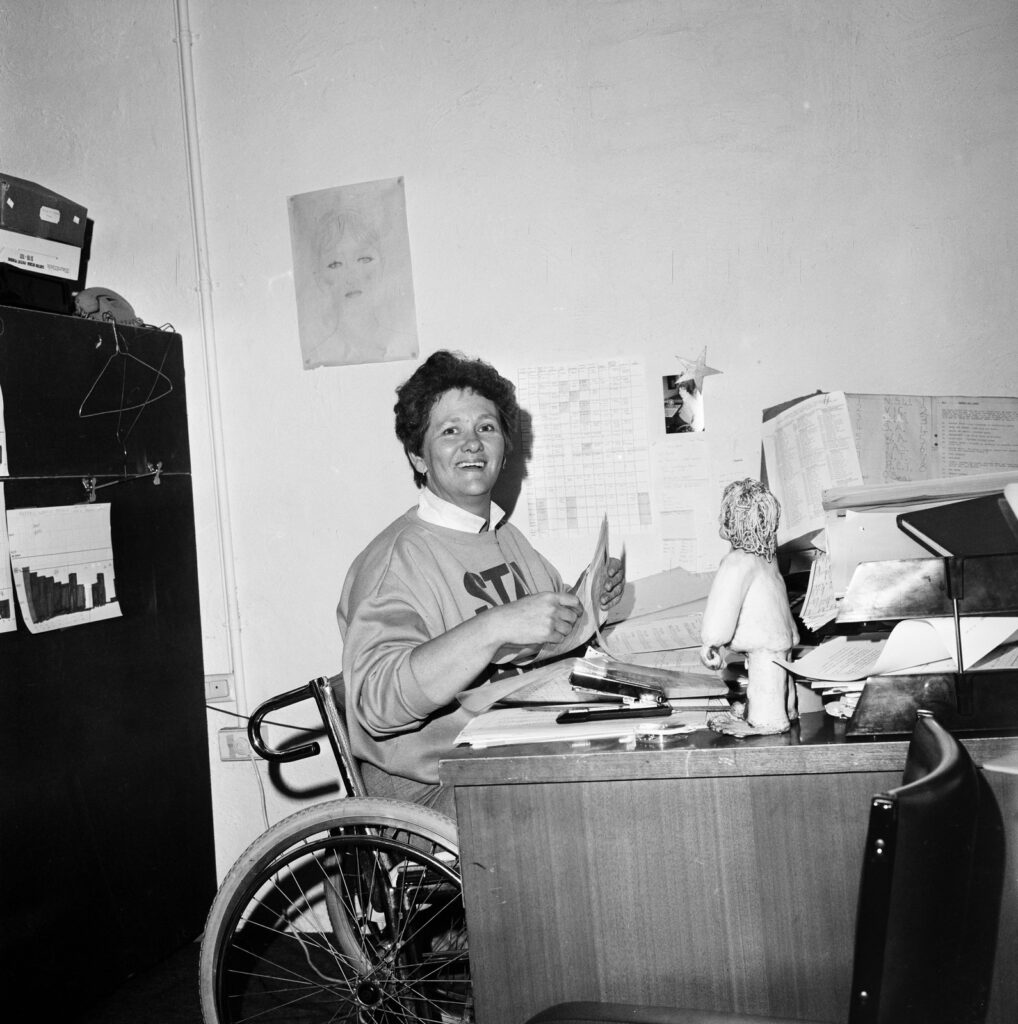Black and white photograph of a woman sitting in a manual wheelchair at a workdesk piled with papers. The woman is smiling, facing the camera.