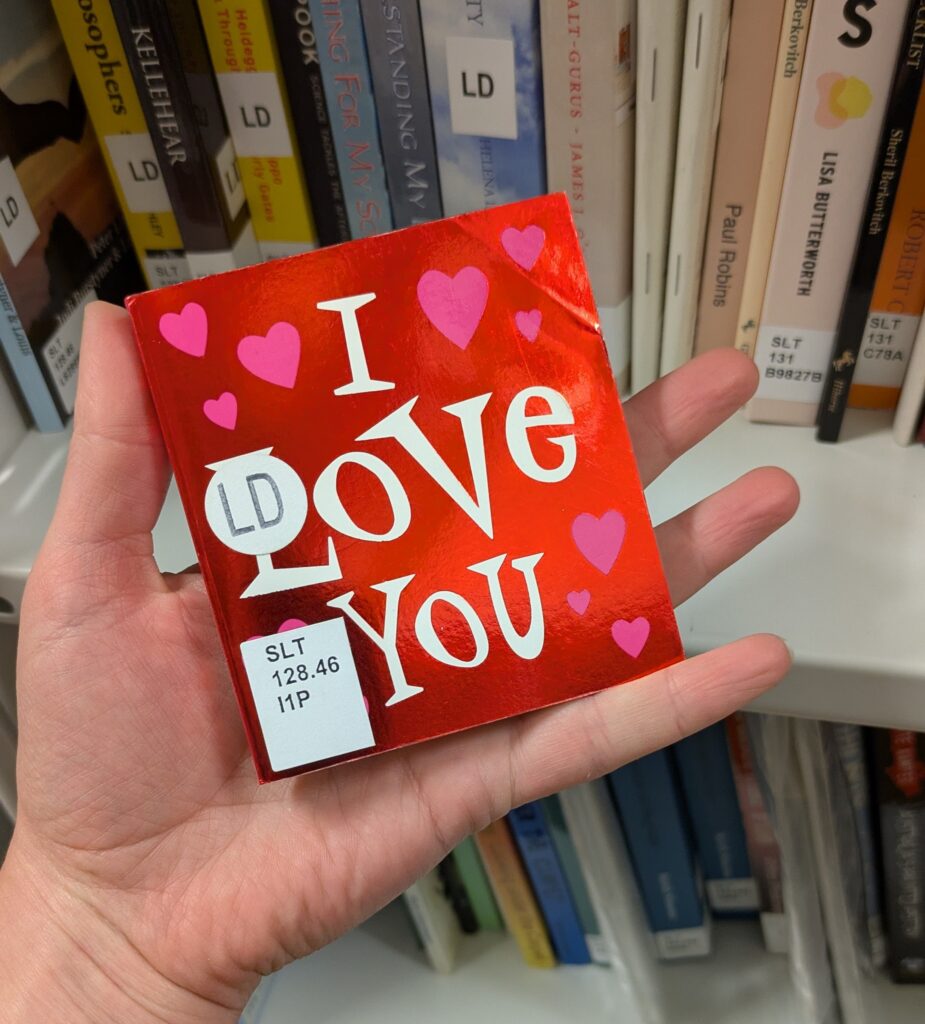 Photograph of a palm-sized shiny red book titled "I Love You", held by a hand in front of a library shelf.