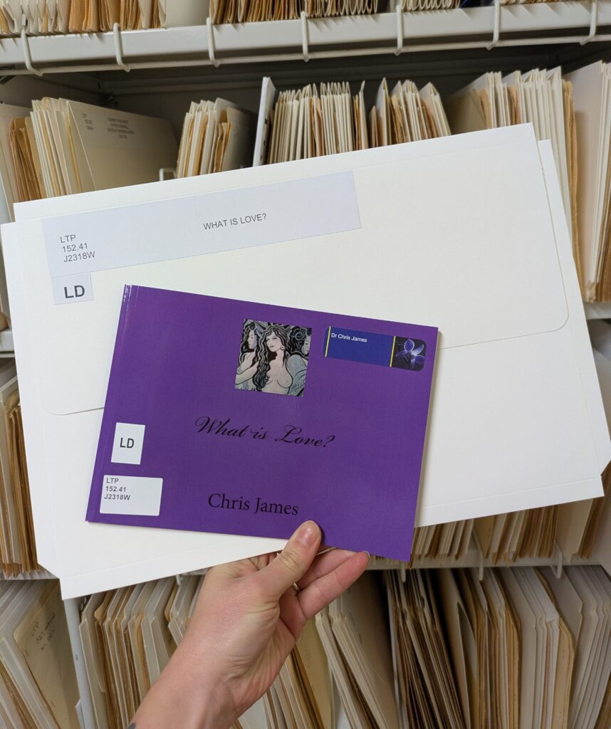 Photograph of a hand holding a purple pamphlet and a white folder in front of a library shelf full of folders. 
