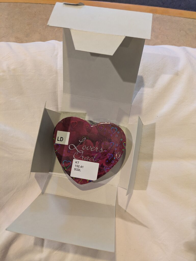 Photograph of an open archival box sitting on a pillow, revealing a heart-shaped tin inside.