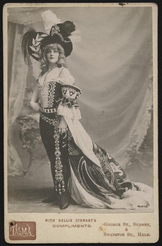 Full portrait of a woman in a velvet and jewelled outfit with a large feathered hat.  Image of actress Nellie Stewart, includes the printed text "with Nellie Stewart's compliments" and a stamp for the Talma photography studio.
