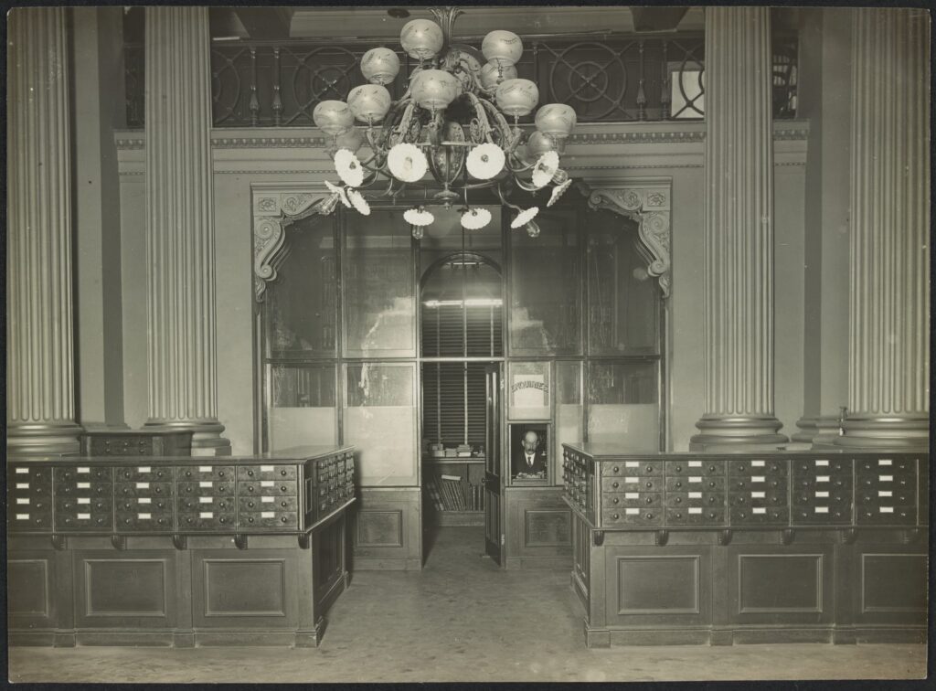 Shows the drawers of the card catalogue and the Enquiries room, built into an alcove. A member of staff sits at the window with "Enquiries" painted on it. A large gasolier, adapted for electricity hangs above the alcove.