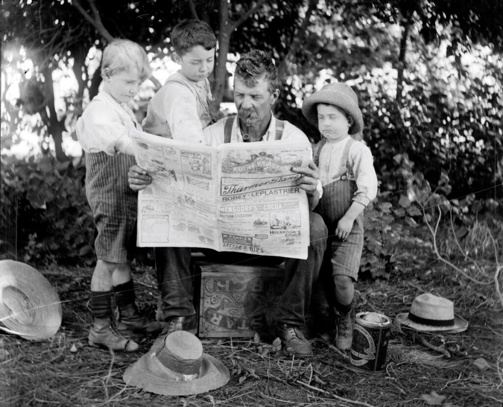 Black and white photo depicts 
man seated on an upturned 'Silver Star Starch' box, smoking a pipe. There are two boys standing on his right and one on his left, all looking at the newspaper. They are outdoors, in a garden. There are hats lying on the ground around them.