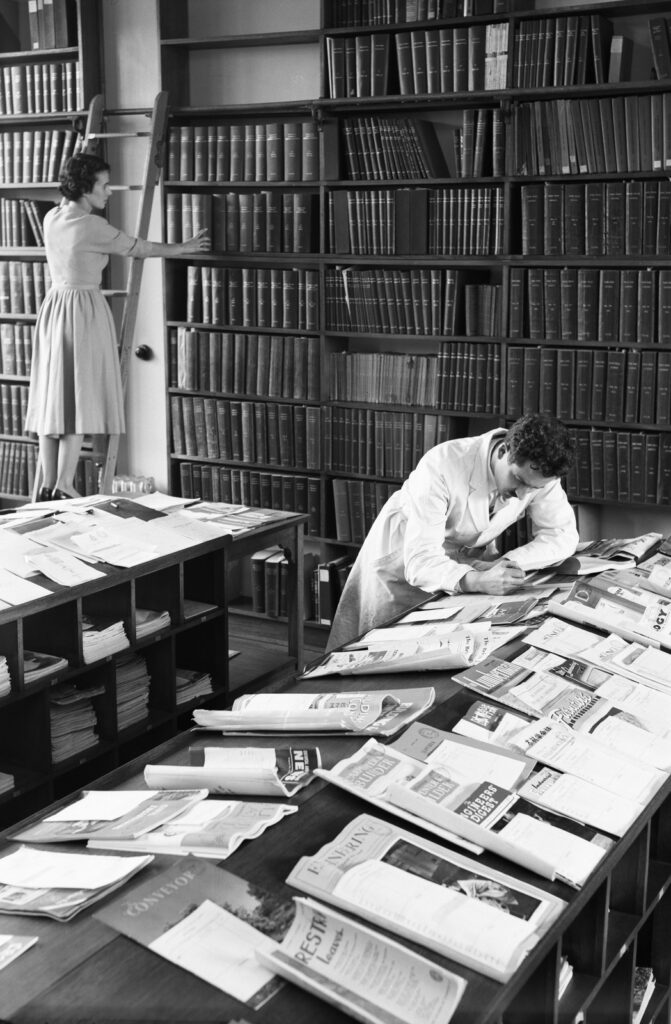 View of a reading room with a female staff member on a ladder shelving a book in background, and a male in the foreground, reading one of several newspapers laid in rows on benches.