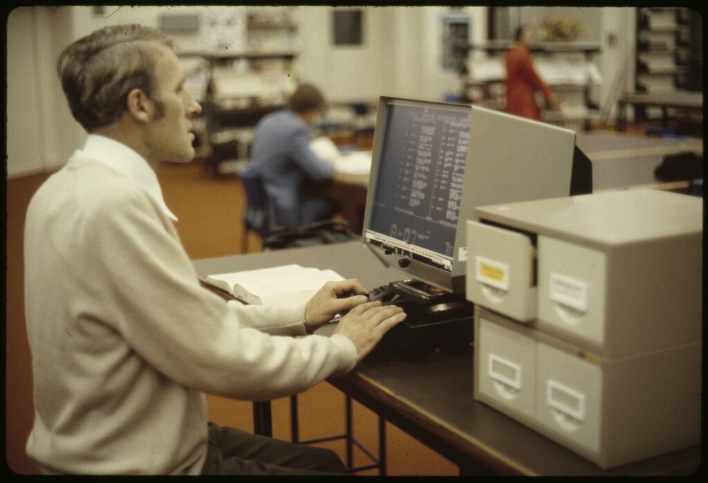 A male seated at a desk, using a microfiche machine, viewing text on the screen.
