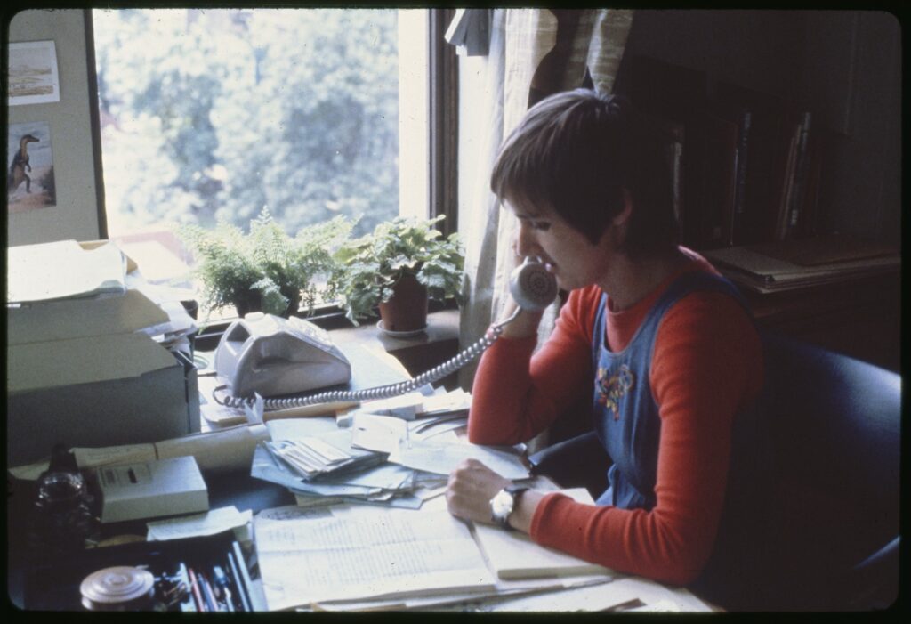 A female staff member with short hair (presumably a librarian) sitting at a desk speaking on the phone, with papers and notes laid out on the desk.
