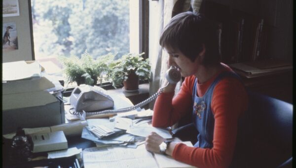 A female staff member with short hair (presumably a librarian) sitting at a desk speaking on the phone, with papers and notes laid out on the desk.