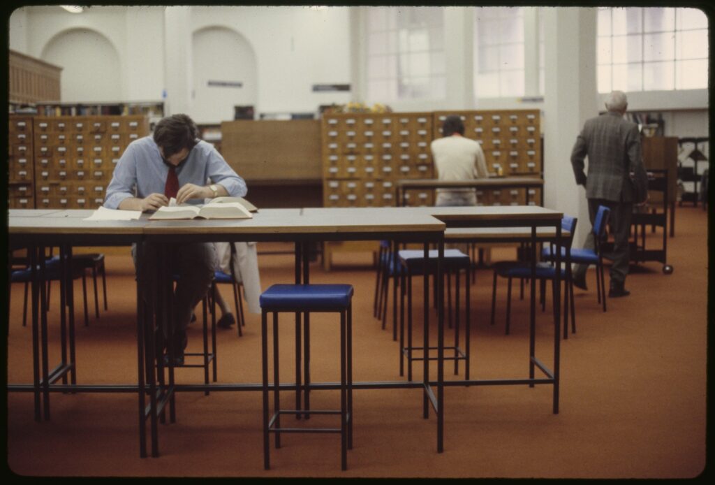View of reading room, dominated by wooden card catalogue drawers. In foreground is a man, sitting at a high bench, reading books.