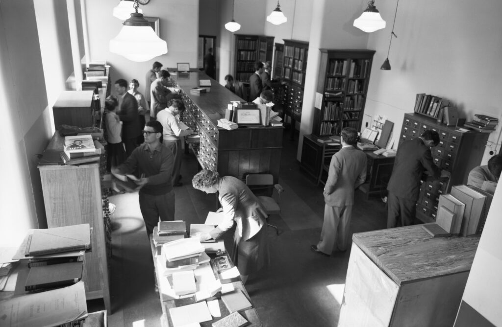A view of reference area, looking down on several people consulting books and card catalogue drawers. There appears to be too many people for a fairly small space. The room is busy. 