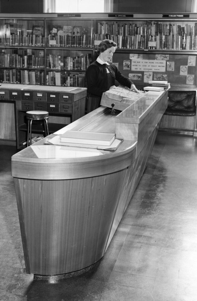 A female staff member (presumably a librarian) standing behind a large reference desk in the library. Shelves of books visible in background, as well as wooden card catalogue drawers.