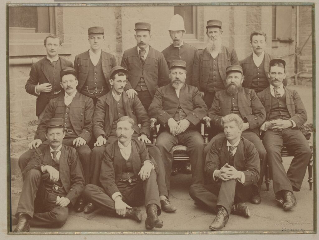 Group portrait photograph of the State Library of Victoria staff. They form three rows, two seated and one standing, and all but two wear a uniform.
