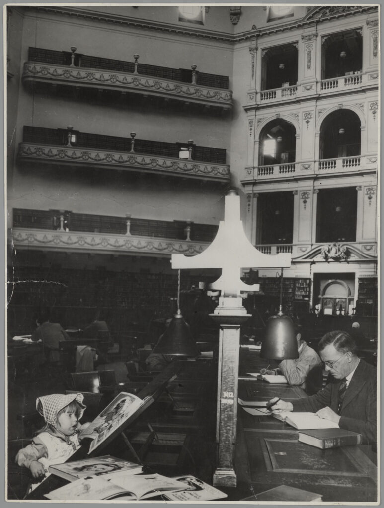Black and white photograph of domed reading room, child with a book on one side of the desk, and an older man reading a book on the other.